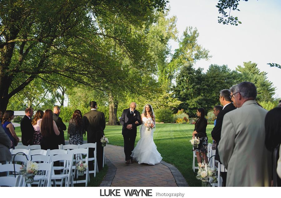 Bride walks down a brick aisle with her father at an outdoor wedding. Guests line the sides.