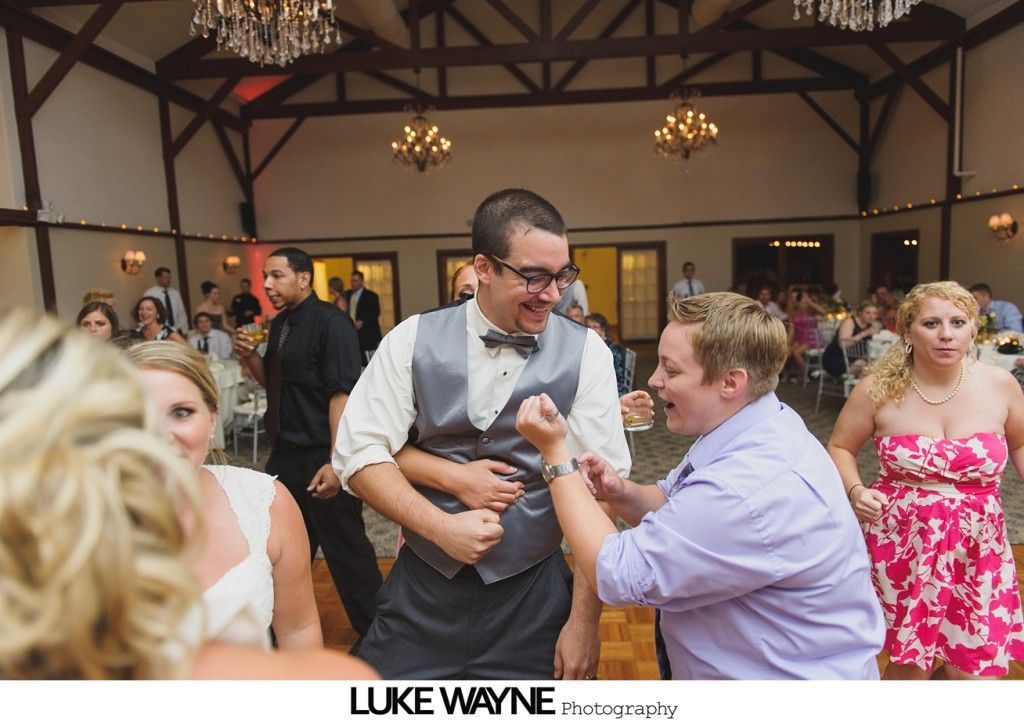 People dancing at a wedding reception. Two men are interacting joyfully, others are in the background, warm interior setting.