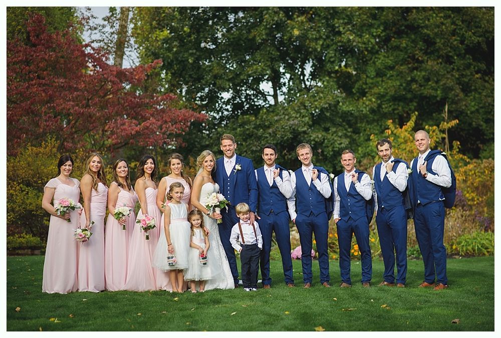 Wedding party poses on green lawn; bridesmaids in pink, groomsmen in blue suits, bride and groom in the center.