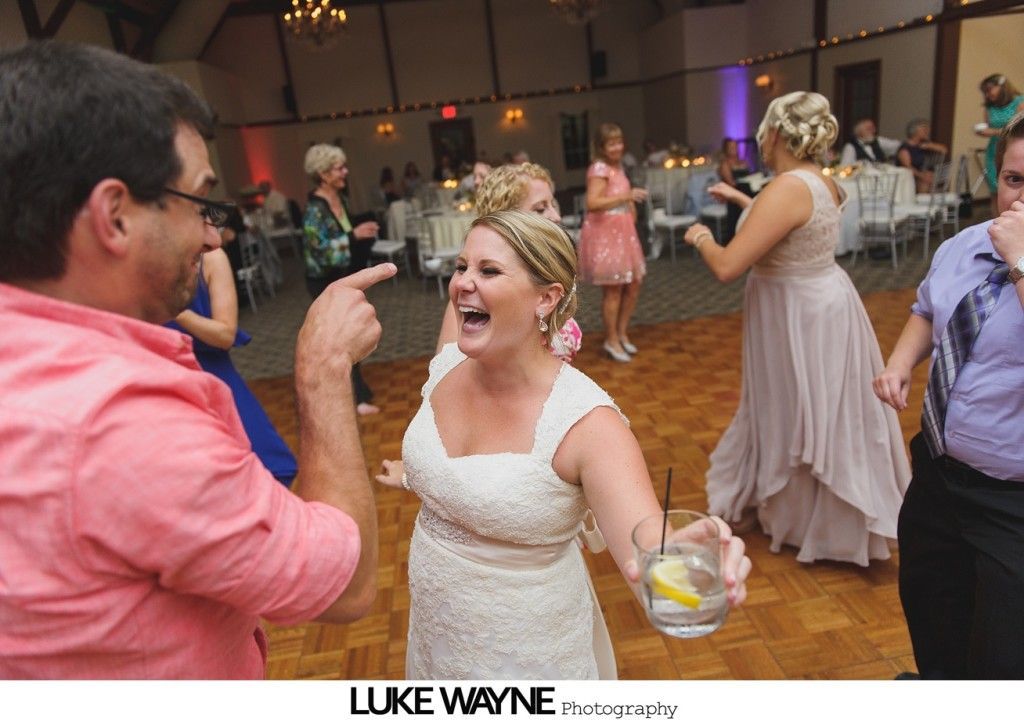 Bride laughing, holding drink, dancing with guests at a reception, others watching.