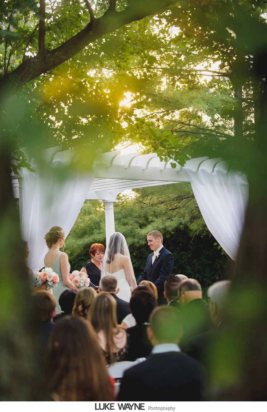 Wedding ceremony under white arbor, with couple, officiant, and guests. Soft sunlight through trees.