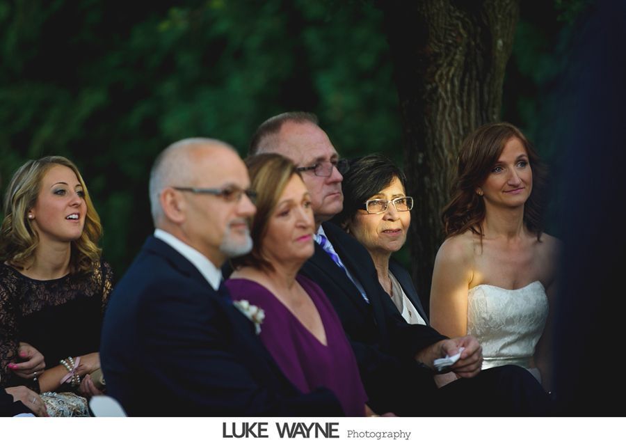 People seated outdoors, watching a ceremony. Woman in a white strapless dress, others in formal wear.