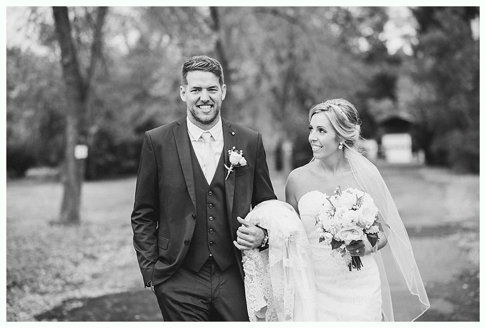 Newlyweds walking arm-in-arm outside; groom in suit smiles; bride in strapless gown holds bouquet.
