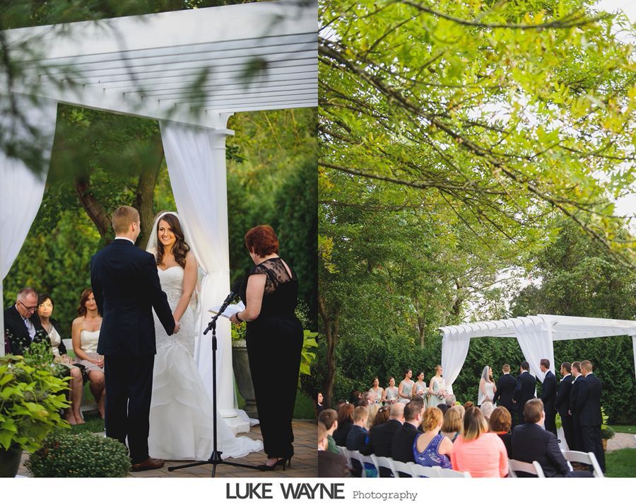 Wedding ceremony outdoors; couple under white arbor, holding hands. Guests seated, officiant in black dress.
