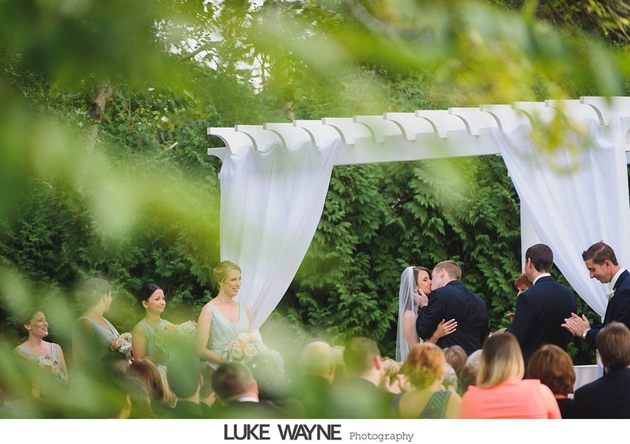 Wedding ceremony: couple kissing under white arch, surrounded by guests in a garden setting.