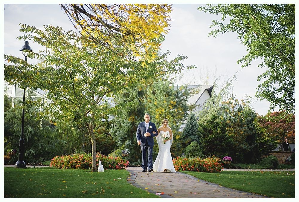 A bride in a white gown and her father walk down a path in a park, sunlight filters through the trees.