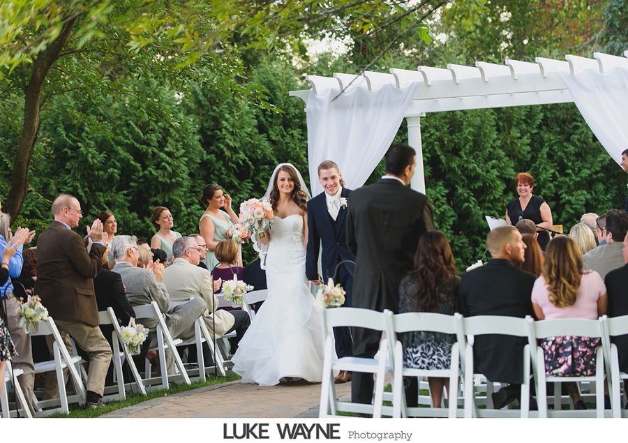 Bride and groom walk down aisle after wedding ceremony, white chairs, green backdrop.