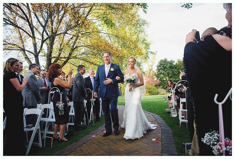 Bride walks down aisle with father, outdoor wedding, guests watching.