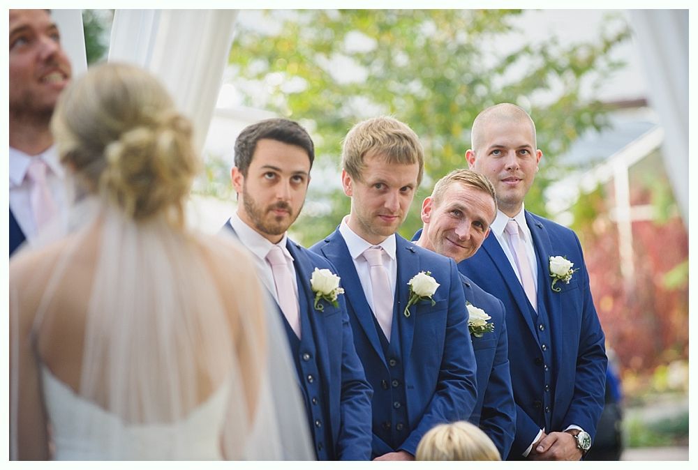 Bride facing away, groomsmen in blue suits, pink ties, and white boutonnieres, looking at her with various expressions during outdoor wedding ceremony.