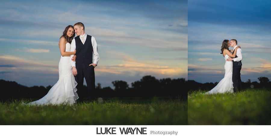 Wedding couple embraces on a grassy hill, sunset sky in background.