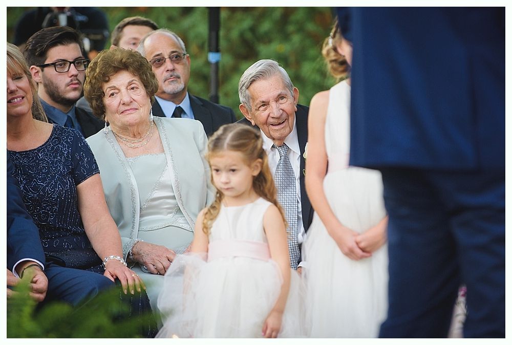 Guests seated at a wedding ceremony, looking toward the couple. People in formal attire. Flower girl in the foreground.