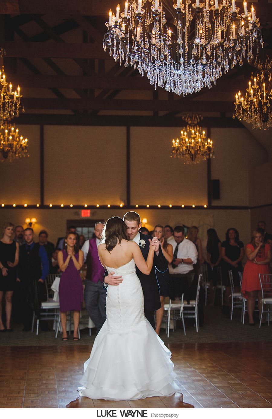 Bride and groom dance under chandeliers at a wedding reception. Guests watch, some clapping.