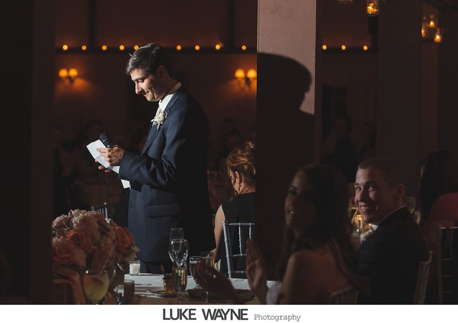 Man in suit giving a speech at a wedding reception; guests smile and clap. Dim lighting.