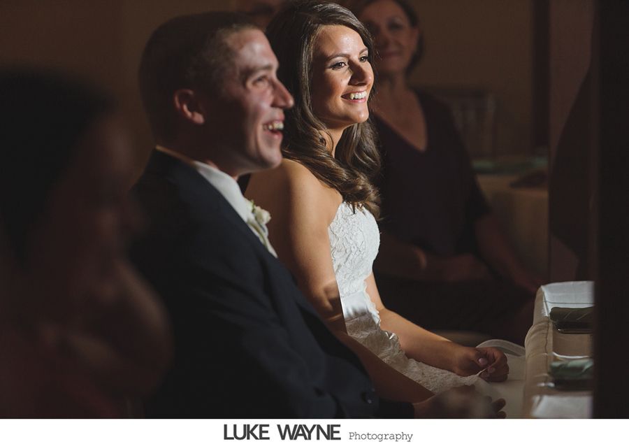 Wedding couple smiling at a reception, dark setting, warm lighting.
