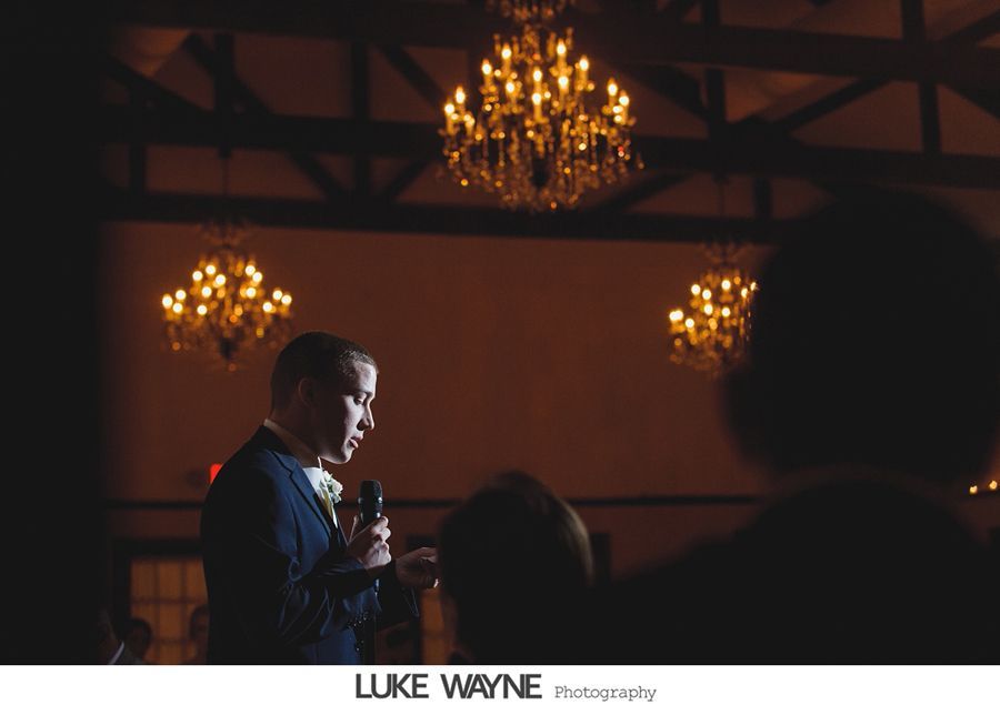 Man speaking into a microphone, illuminated by overhead chandeliers in a dark room.