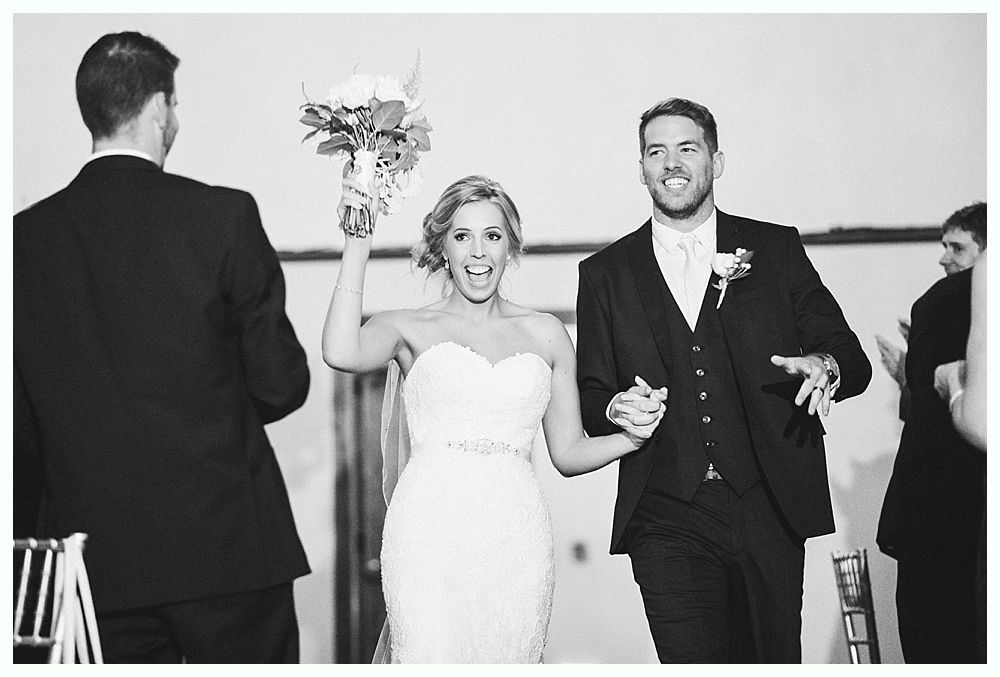Newlyweds celebrate, walking with excitement, holding hands, woman raising bouquet. Black and white.