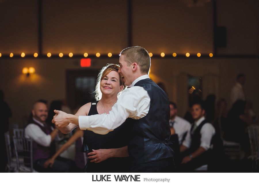 Groom dancing with woman in a dark dress at wedding reception. Others watch. Warm lighting.