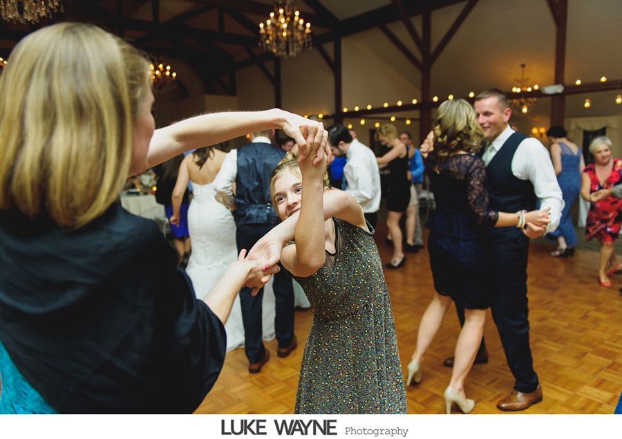 People dancing at a wedding reception on a wooden dance floor, with chandeliers hanging overhead.
