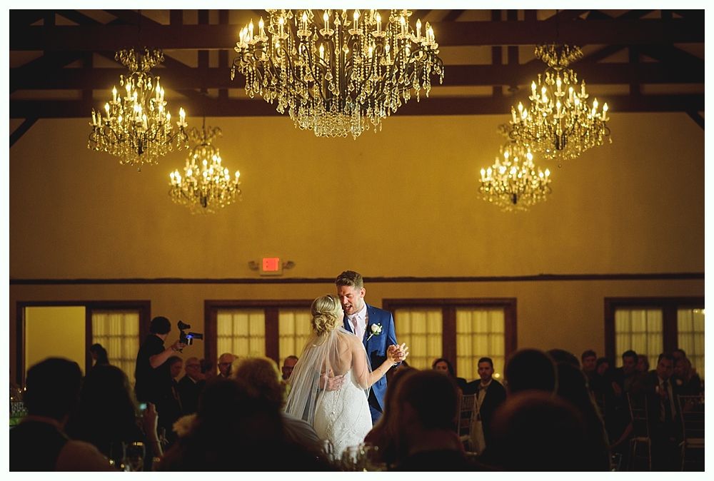Couple dancing at a wedding reception under chandeliers; guests watching.