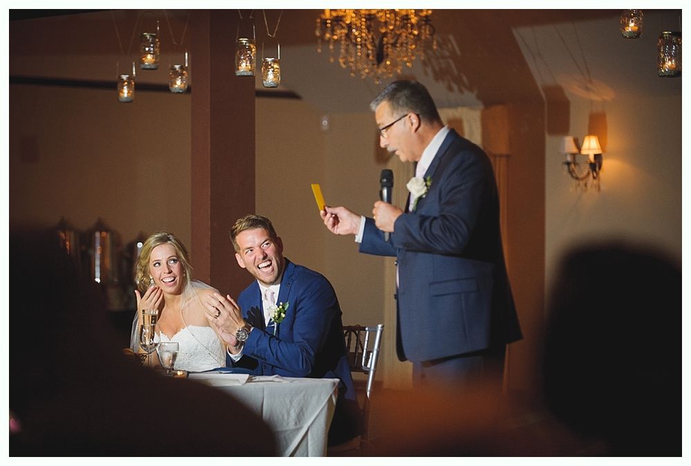Man giving speech at wedding; couple reacts, bride smiles, groom laughs. Indoor setting, chandelier, neutral tones.