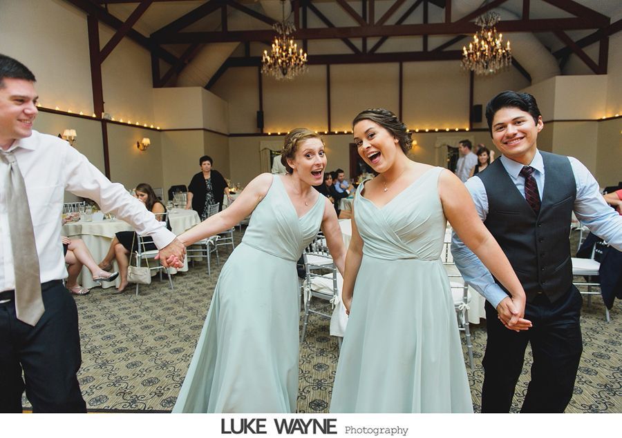 People holding hands, dancing at a wedding reception. Two women in teal dresses, one man in a vest, one in a white shirt.