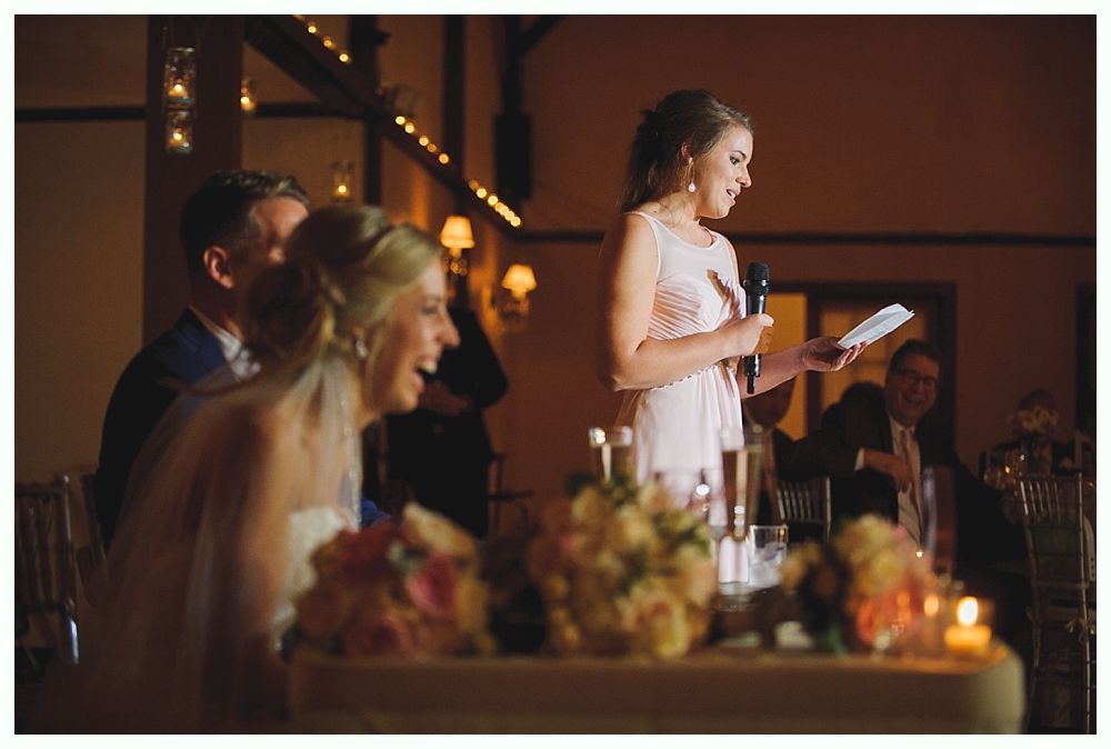 Woman giving a speech at a wedding, bride and groom laugh. Dimly lit room with floral arrangements.