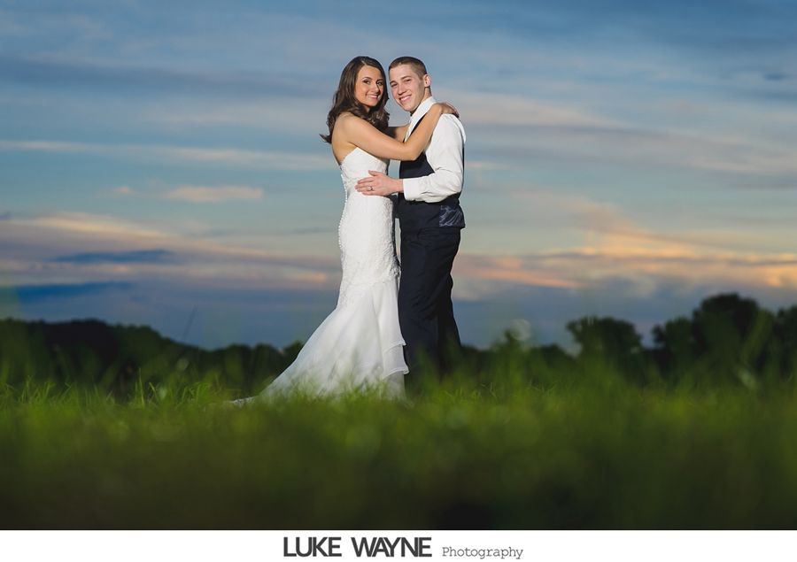 Bride and groom embracing in a field at sunset, wearing wedding attire.