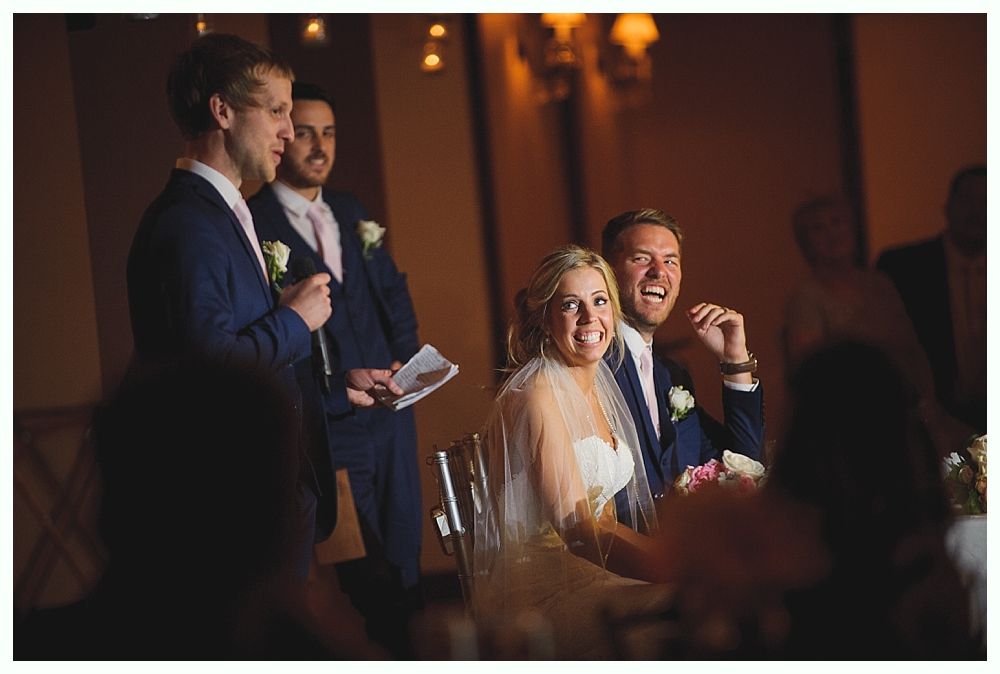 Bride and groom laugh at a wedding toast; groomsmen in suits stand nearby.