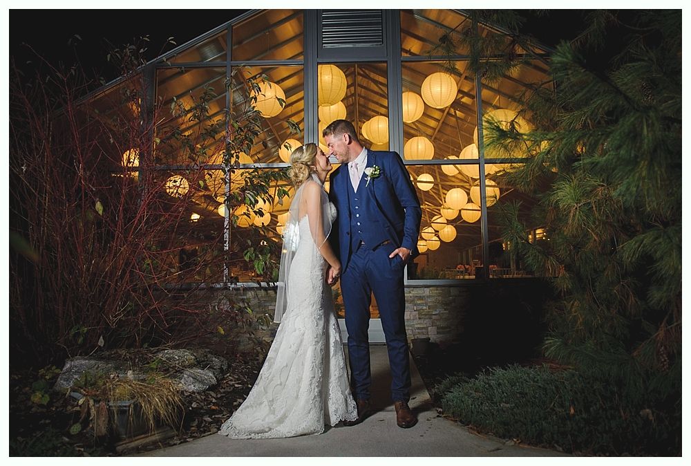 Newlyweds kissing in front of a glass building adorned with round lanterns at night.