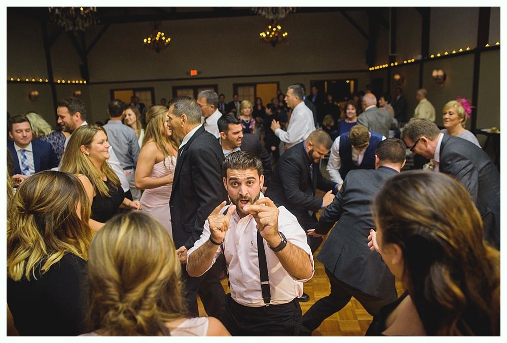 People dancing at a wedding reception; man in foreground making finger guns, everyone smiling. Wooden dance floor in a hall.