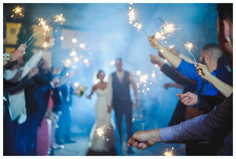 Newlyweds walk through sparkler-lit tunnel, guests holding sparklers, blue-toned night scene.