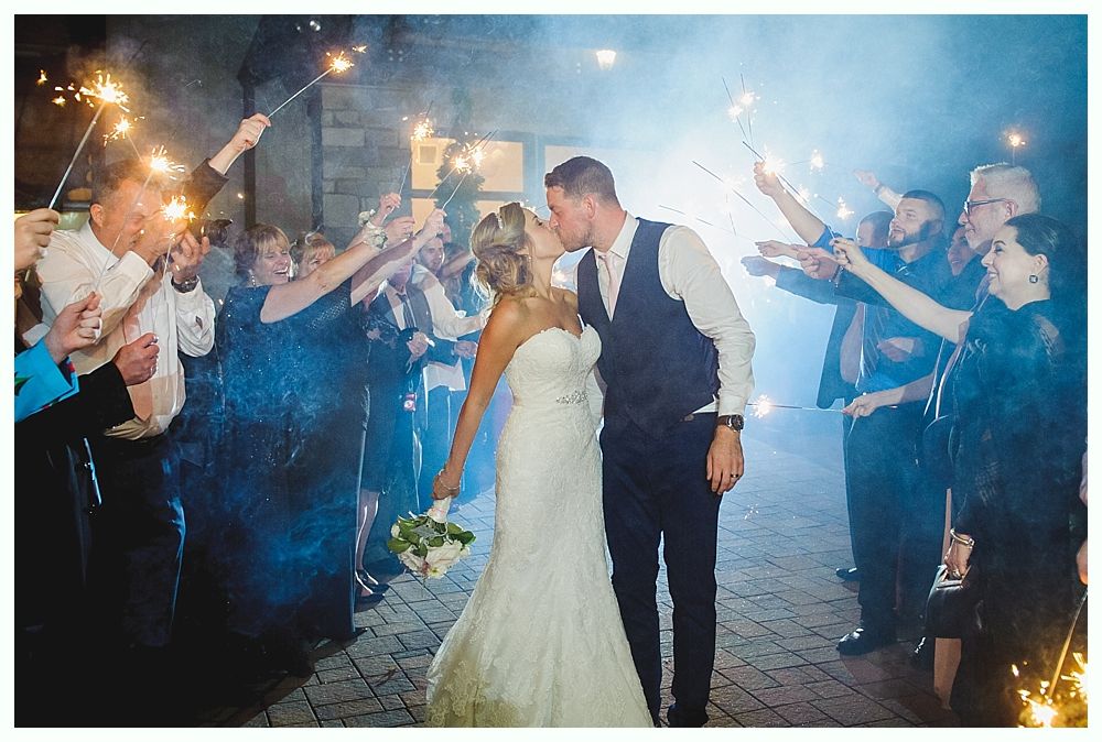 Bride and groom kiss as guests hold sparklers at a wedding, night setting.