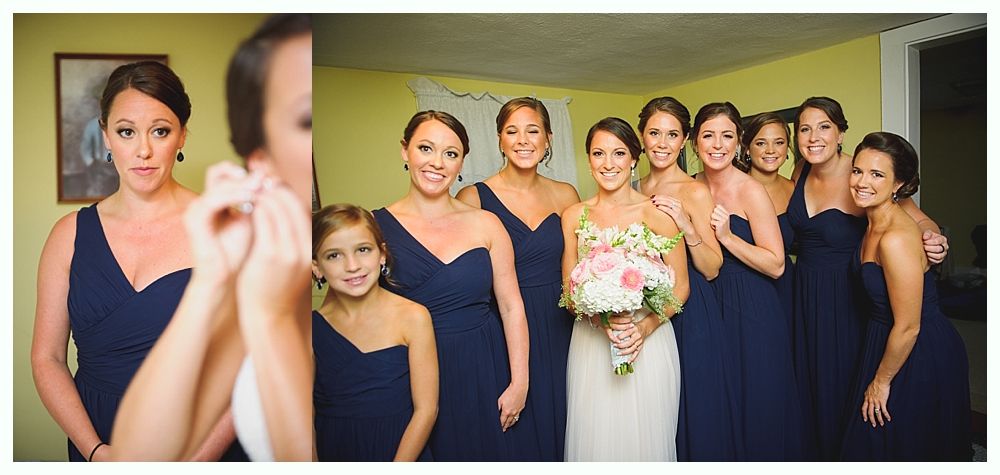 A bride and bridesmaids in navy dresses pose, some smiling, some touching their ears, in a room.