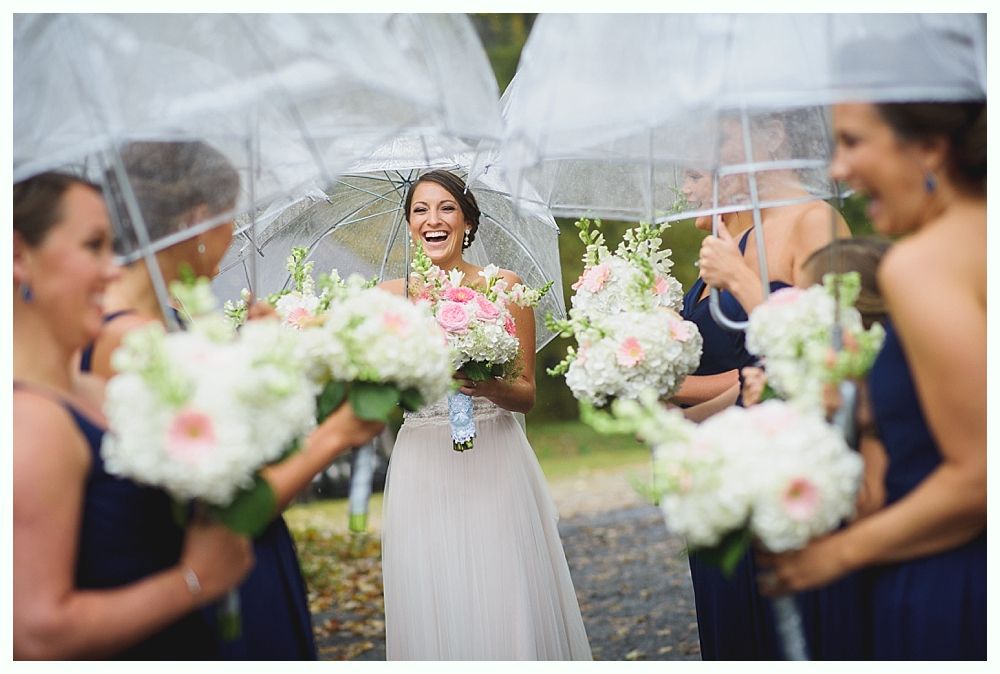 Bride laughs with bridesmaids under umbrellas in the rain, holding bouquets.