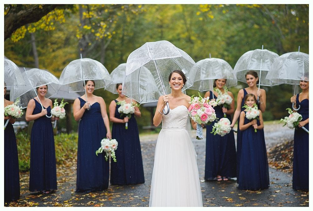 Bride and bridesmaids in navy dresses hold bouquets and clear umbrellas outdoors in the rain.