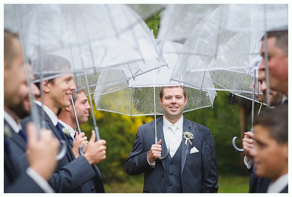 Groom smiles, surrounded by groomsmen with clear umbrellas in the rain. They are in a garden.