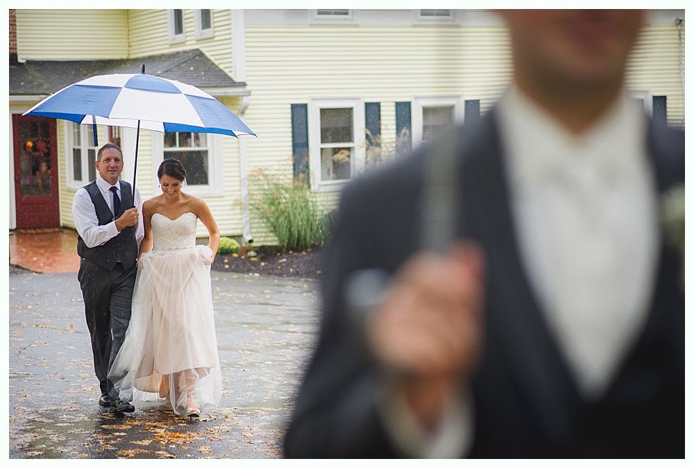 Bride and man with umbrella walk in rain, next to a yellow building.