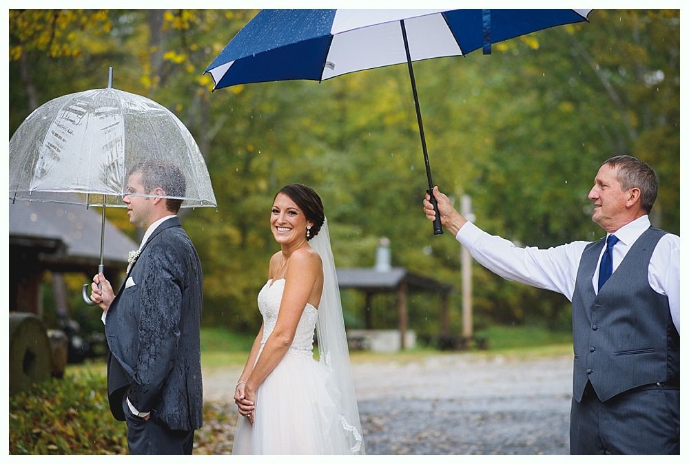 Bride and groom embrace, one kissing. Outdoor wedding setting with trees and flowers. The couple smiles joyfully.