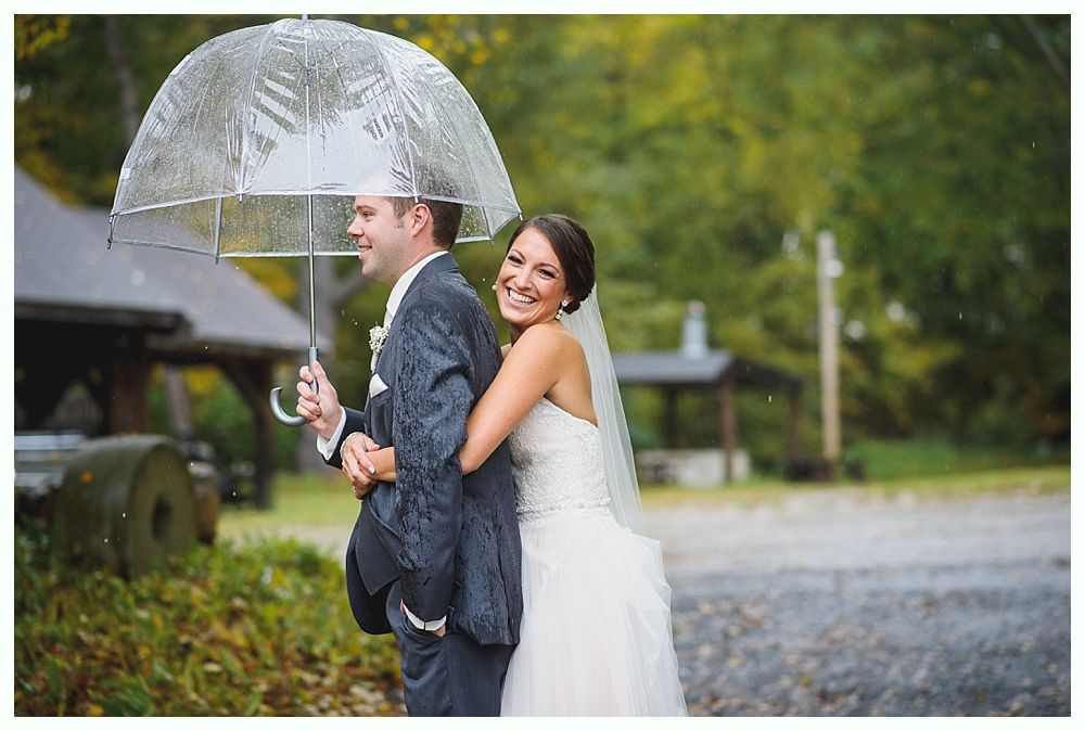 Wedding couple under clear umbrella, bride hugging groom, smiling outdoors.