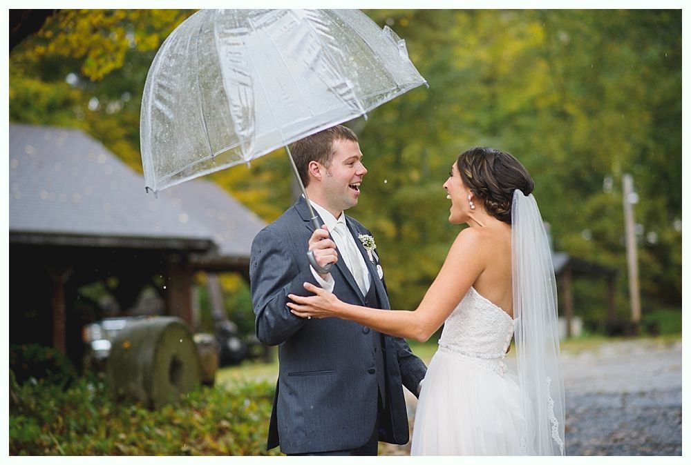 Couple laughing under clear umbrella on their wedding day; outdoor setting.