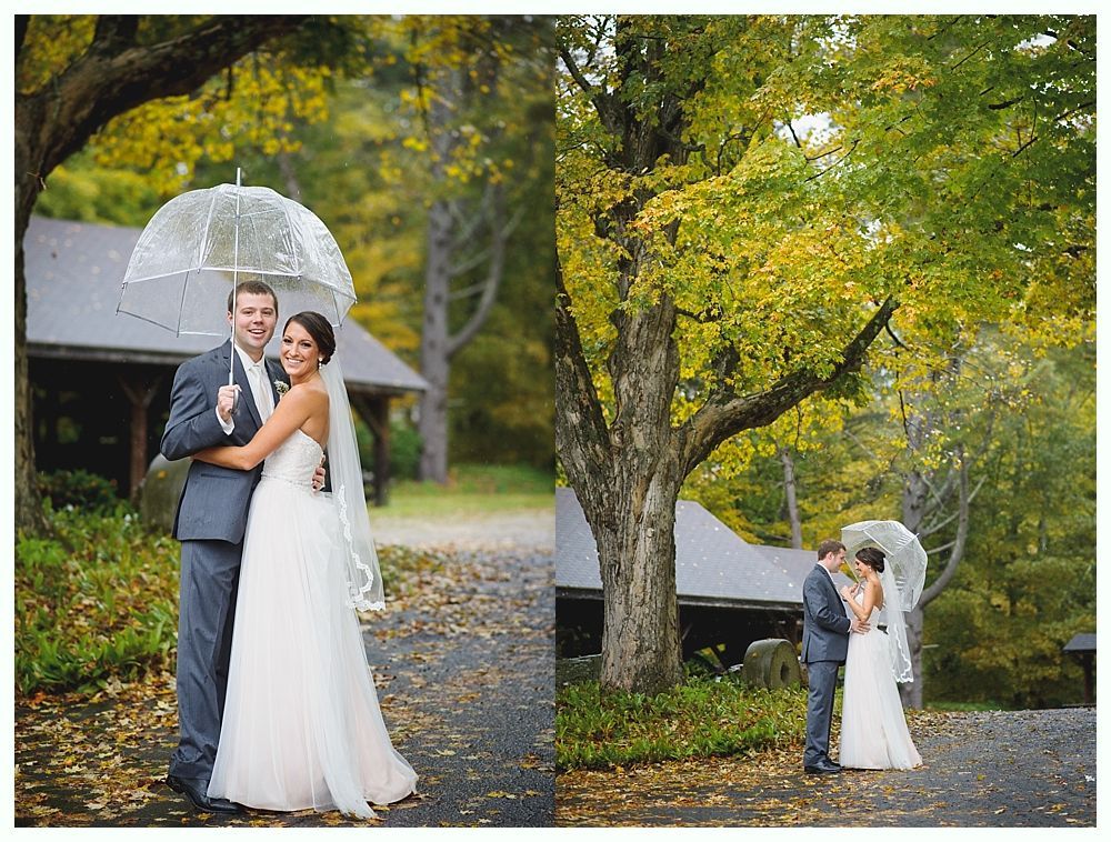 Bride and groom embrace outdoors, holding umbrella. Autumn leaves and trees surround them.