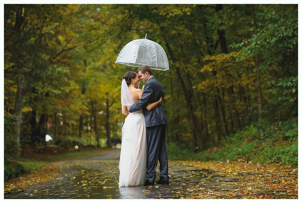 Couple embraces under an umbrella on a rainy day. Forest backdrop, bride in white dress, groom in a suit.