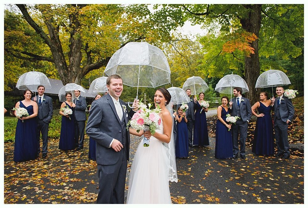 Wedding party under clear umbrellas in a park, bride and groom laughing. Others in navy dresses and gray suits.