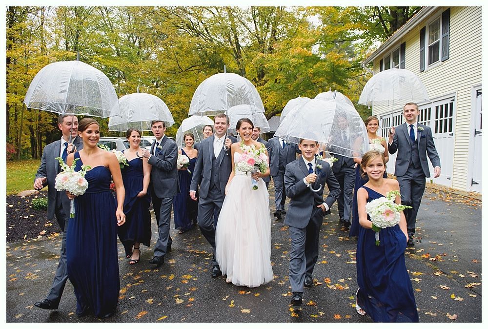 Wedding party walking under clear umbrellas in the rain. Bridesmaids in navy, groomsmen in gray, smiling.