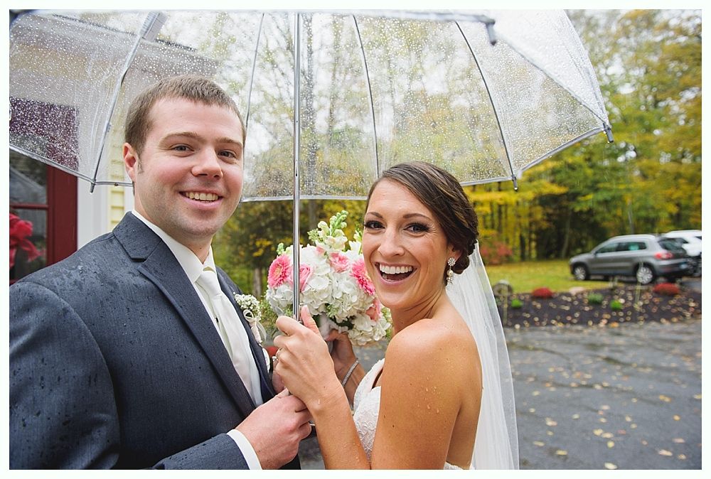 Bride and groom under a clear umbrella smiling on wedding day; bouquet in hand, outdoors.