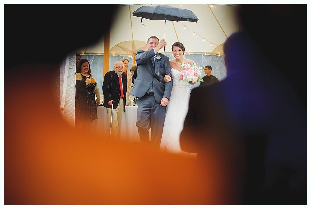Newlyweds exiting ceremony under an umbrella, smiling. Guests watching. Rainy outdoor wedding.