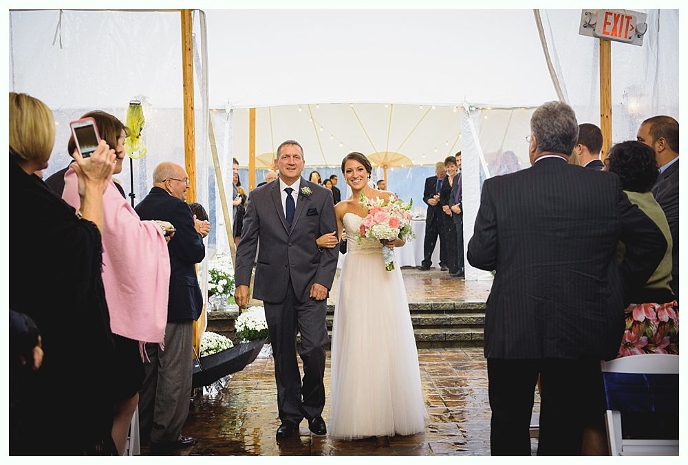 Bride and escort walk down the aisle at an outdoor wedding. She carries a bouquet; guests watch.