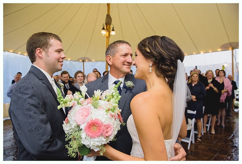 Wedding ceremony: Bride and groom facing each other, father smiling between them. Guests watch in tent.