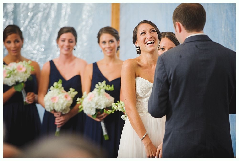 Bride laughing during a wedding ceremony, bridesmaids in navy dresses holding bouquets; groom faces her.