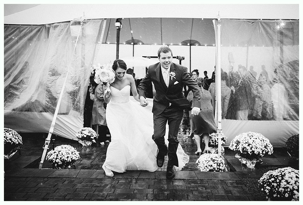 Bride and groom exiting wedding reception tent, smiling, holding hands, with floral decorations.